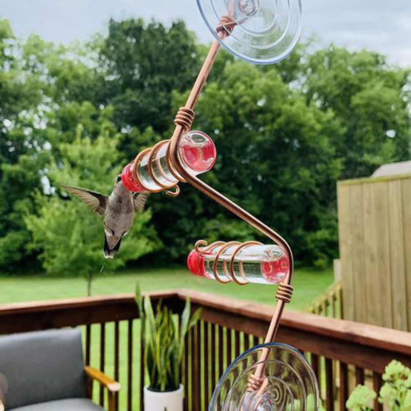 Window hummingbird feeder with copper wire frame and red cups, hummingbird in flight